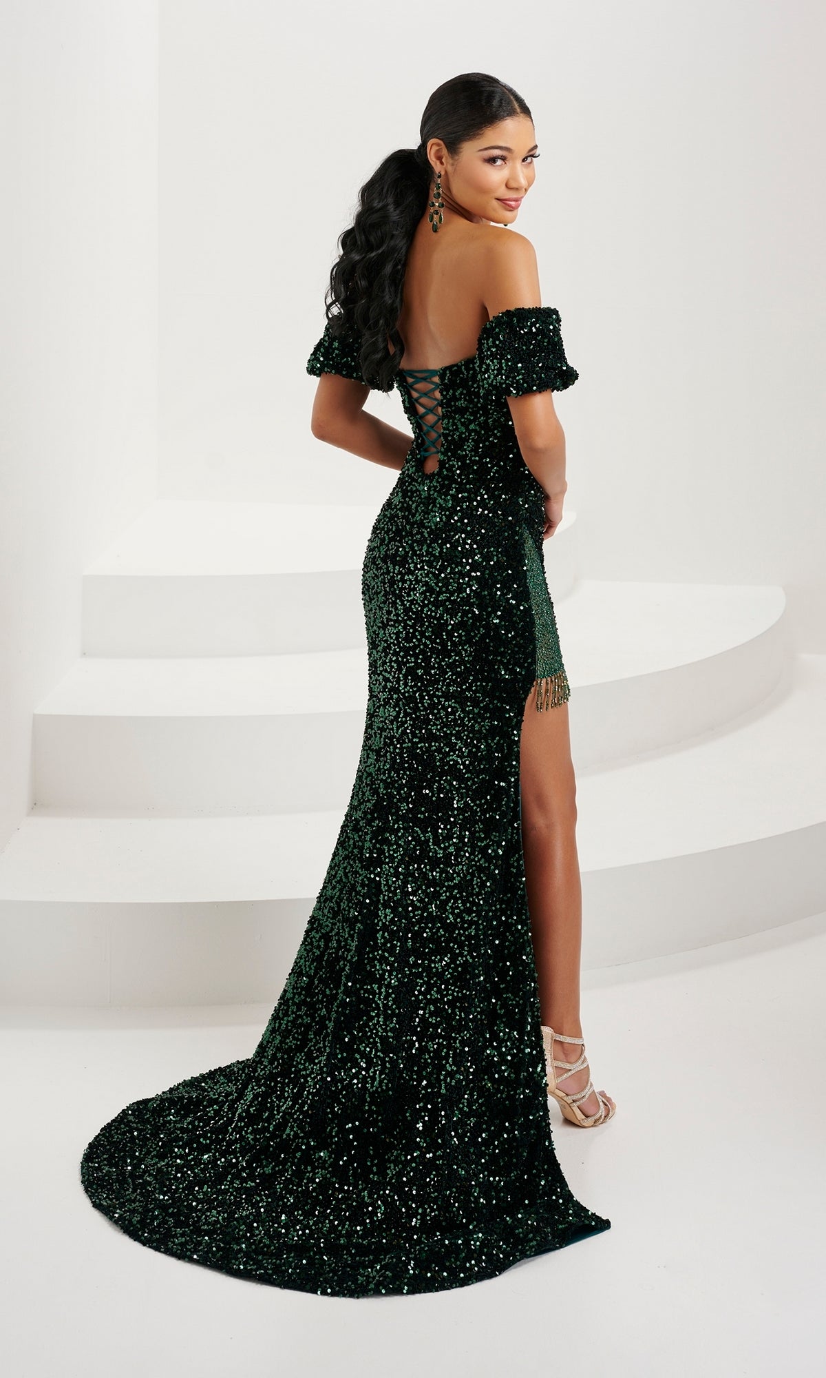 A woman in the Tiffany Navy Blue Sequin Velvet Prom Dress 16087—featuring an off-shoulder design, lace-up open back, and thigh-high slit—stands on white steps, glancing over her shoulder.